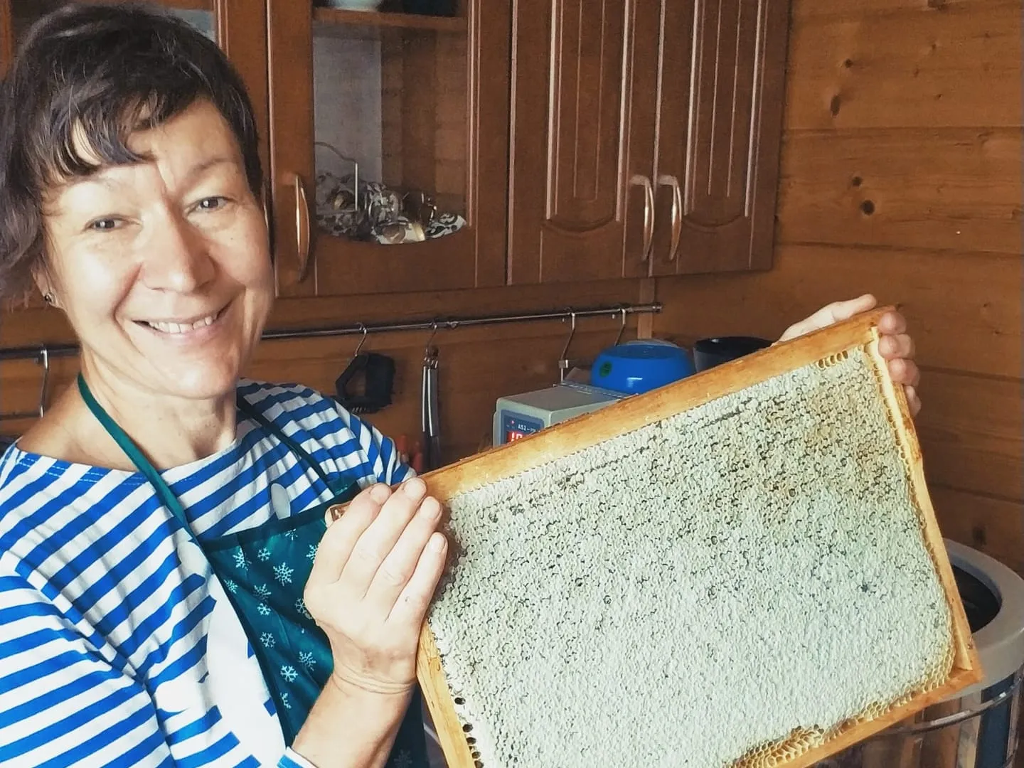 Пасечница в полосатой тельняшке и фартуке держит рамку с запечатанными сотами; за спиной деревянная кухня, рядом медогонка